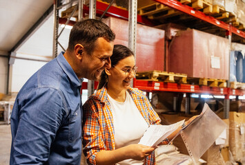 Smiling woman and man in casual clothes are working in warehouse. Young couple of employees laugh and mark presence of product storage in workshop.