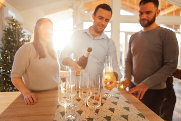 Smiling young man pours champagne into glasses in dining room of house.