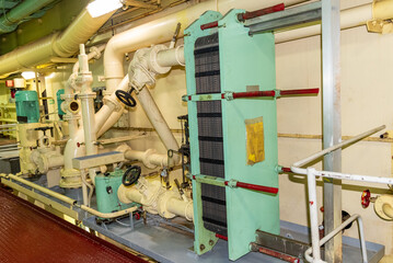 The engine room interior featuring a heat exchanger, oil cooler, and its instrumentation, showcasing the essential equipment for maintaining the ship's mechanical systems.
