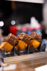 Close-up of a delicious chocolate-covered pastry with sprinkled decorations, displayed on a metal rack in a bakery or cafe setting. Perfect for illustrating gourmet desserts, sweet snacks, and food ph