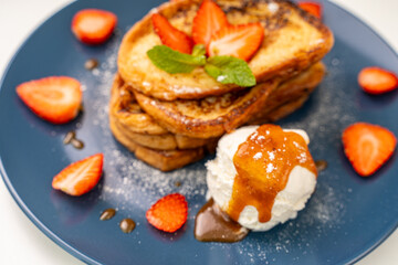French toast decorated with strawberries and mint leaves with ice cream drizzled with caramel sauce and powdered sugar on a blue plate and a glass of orange juice on a white background, table