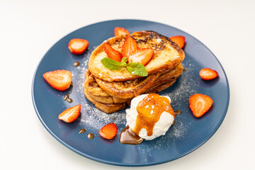 French toast decorated with strawberries and mint leaves with ice cream drizzled with caramel sauce and powdered sugar on a blue plate and a glass of orange juice on a white background, table