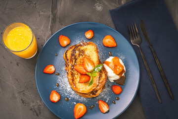 French toast with strawberries and mint, ice cream topped with caramel sauce on a blue plate with cutlery on a napkin and orange juice on a gray background. Healthy breakfast concept.