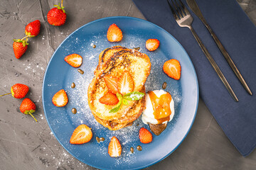 French toast with strawberries and mint, ice cream drizzled with caramel sauce on a blue plate with cutlery on a napkin on a gray background. Healthy breakfast concept.