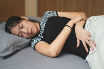 Frustrated young asian woman with menstrual pain and having cramps during period, touching her belly, lying in bed and cannot even sleep through this pain.