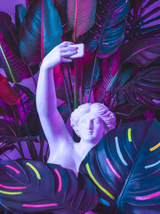 A greek sculpture of Venus or Aphrodite holding her smartphone to make a selfie. She's among tropical vibrant palm and monstera leaves. Some of them are painted.