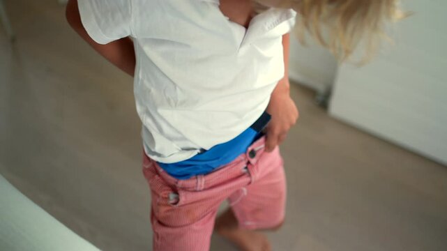 Young boy adjusting and buttoning his pants, focusing on getting dressed while standing in a home environment, viewed from above