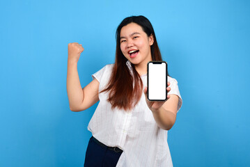 Naklejka premium Smiling young asian woman wearing t-shirt give thumbs up gesture to vertical blank smartphone screen for advertising, isolated over blue background with copyspace