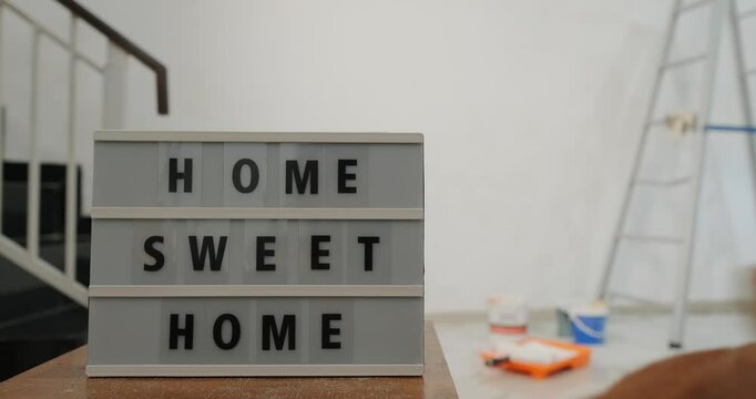 In the foreground is a sign that says Home Sweet Home. In the background is a golden retriever dog running down the stairs, wagging its tail as it runs past the sign. The apartment is being renovated.