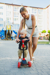 A mother teaches her little son to ride a scooter. Games of mother and child on the street. The concept of a family walk with a child