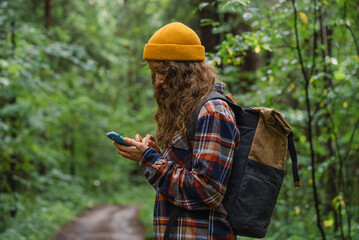 Naklejka premium Woman in a yellow beanie and plaid shirt checks her smartphone while hiking through a lush green forest, carrying a backpack. 