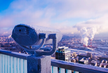 Binoculars coated in frost stand at a high vantage point, offering a view of a city blanketed in...