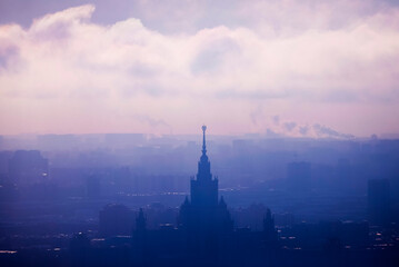 Obraz premium A Moscow State University building in Moscow stands silhouetted against a misty sky at dawn in soft clouds with hints of industrial smoke in the background, capturing the calmness of the morning.