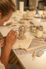 Close-up of an artisan shaping a small clay figurine on a workbench with pottery tools, striped...