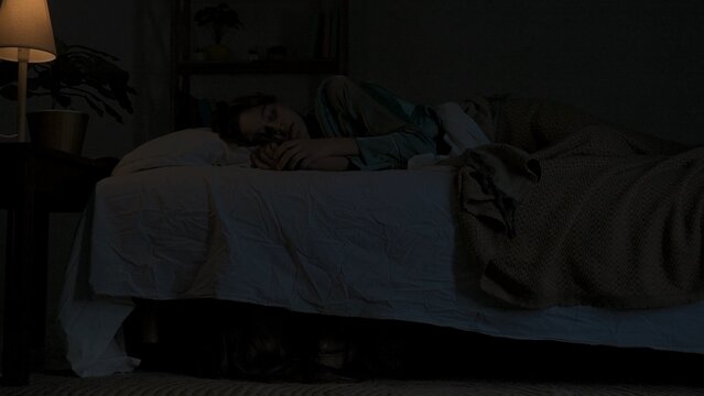 Woman sleeping in bed in dark apartment room, monster hiding under bed. Storm and lightning flash lights outside. Halloween horror concept.