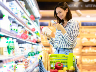 Smiling young woman taking dairy products from shelf in the supermarket, holding bottle and...