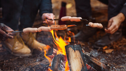 Fototapeta premium Cropped of mixed race group of friends sitting beside fireplace and frying sausages in forest