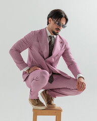 cool fashion guy with sunglasses in pink suit crouching on wooden chair