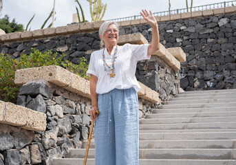 Smiling 70 year old elderly woman with walking disability holding stick knob while walking down stairs outdoors, leaning on stick, suffering from muscle problems, bone disease, recovering from trauma