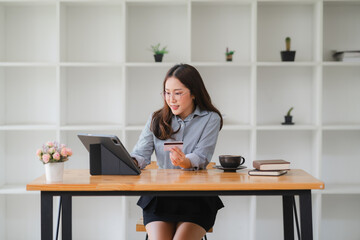 Online Shopping at Home: A smiling young Asian woman sits at her desk, confidently making a...