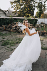 Fototapeta premium Beautiful young curly smiling blonde bride in a white dress outdoors dancing, spinning around herself against the background of logs in nature in the village.
