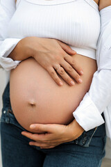 Young pregnant woman, beautiful girl holding big belly with hands in jeans and white shirt standing outdoors. Close-up photo, portrait, pregnancy concept.