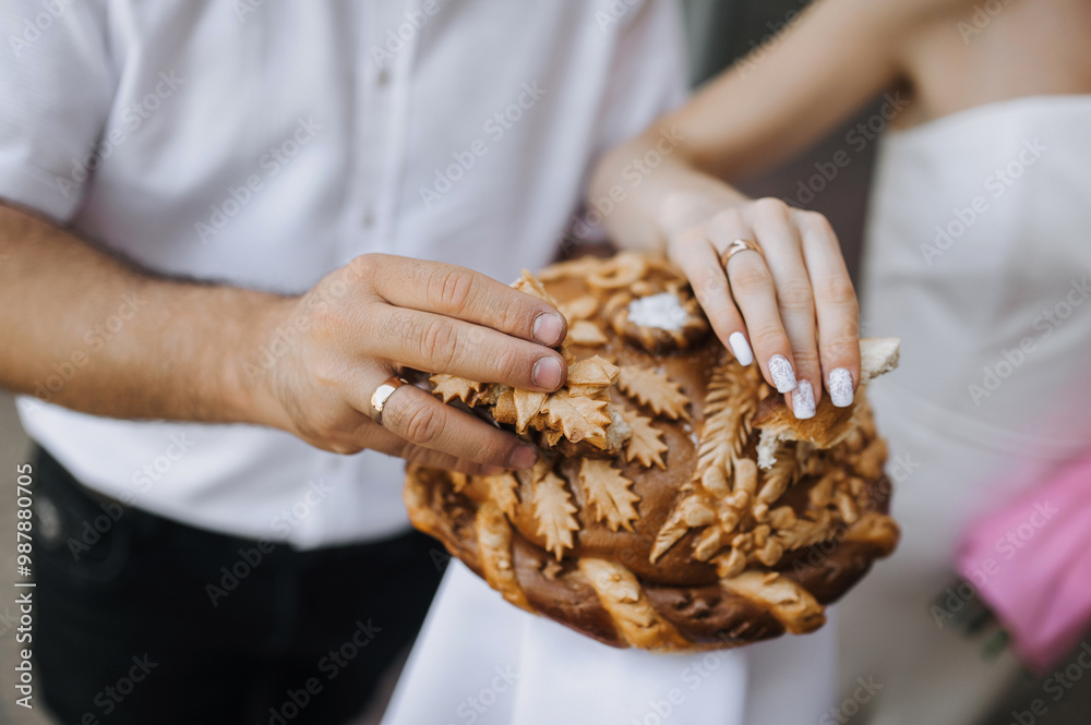 Wall mural Happy bride and groom break off a piece of decorated Ukrainian loaf, fresh pastry with salt and a towel with their hands wearing gold rings on their fingers. Photo of food, tradition at a wedding.