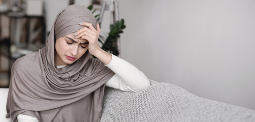 Unhappy muslim woman in headscarf sitting on sofa and touching head in despair, feeling depressed...