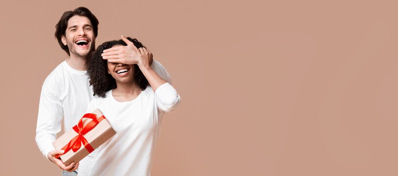 Surprise. Loving man prepared romantic present to his girlfriend, holding gift box and covering her eyes over yellow background in studio, empty space