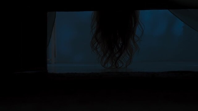 Woman on the bed in dark room looking down underneath the bed for monster, scares and hides away, view from underneath bed. Halloween horror concept.