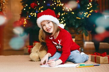 Xmas smiling girl sitting next to festive tree and drawing, looking at camera, copy space
