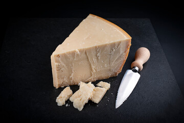 Traditional Parmesan cheese piece with cheese knife during food preparation as close-up on a black design board with text space