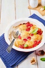 Fish meatballs baked with tomatoes and sauce in a ceramic baking dish on a white wooden background.