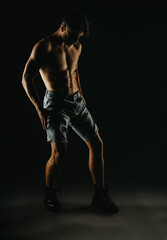 Young athletic man in denim shorts showcasing muscular physique in a dimly lit studio with dramatic shadows.