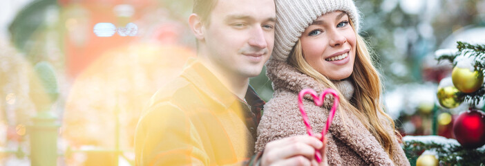 Beautiful young loving couple, boyfriend and girlfriend having fun on a Christmas market wearing warm clothes, hat. Outdoors, winter time, snowy weather. Bright candy canes in heart shape. Banner