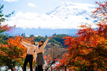 Happy Asian woman travel Japan Fuji on holiday vacation. Attractive girl friends using mobile phone taking selfie during travel Mt.Fuji and looking beautiful red maple tree leaf falling down in autumn