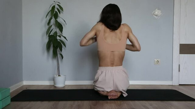 Beautiful fit atletic body woman doing stratching exersices for shoulders, scapula and healthy back in bra and sporty wear sitting on black mat at home interior. Health yoga and sport lifestyle