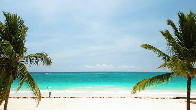 Palm trees frame view, creating perfect atmosphere for relaxation. In background, few people walking along shore. There ample space for text, making it perfect for advertising or promotional material.