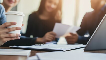 Close-up of young business professionals collaborating on a laptop in an office, focusing on their hands with documents and coffee, while a team discusses a project in the background
