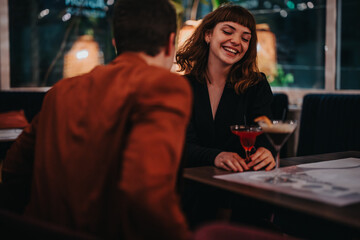 A young couple smiles and laughs during a night out at a restaurant. They enjoy cocktails and good conversation in a cozy, intimate setting.