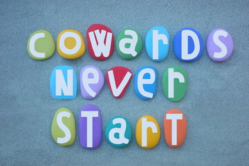 Cowards never start, motivational slogan design composed with hand painted multi colored stone letters over green sand