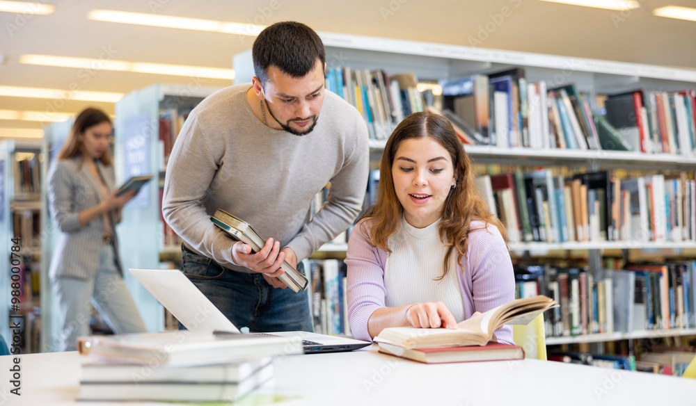 Canvas Prints Two friendly young adult male and female students preparing for exam together in university library, using textbooks and laptop