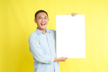 Asian man in light blue denim shirt holding blank white board for advertisement isolated on yellow...
