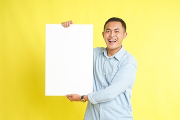 Asian man in light blue denim shirt holding blank white board for advertisement isolated on yellow background