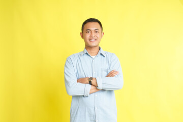 confident smiling Asian man in light blue denim shirt with arm folded isolated on yellow background