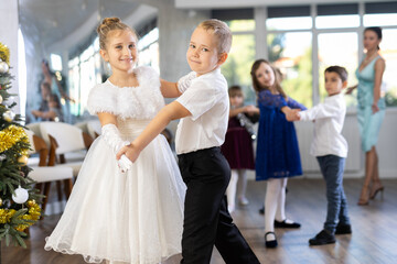 Fototapeta premium Enthusiastic preteen children, girls and boys in elegant outfits practicing partner dance moves in studio adorned with Christmas decorations, under guidance of female teacher