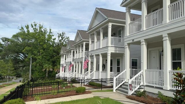 Tallahassee, FL USA - July 1, 2024: Handheld medium-shot video of American flags hung over the porches of white row houses. The shot slowly pans to the right to reveal more of the houses. 