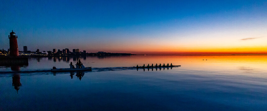 A rowing team glides across calm water at sunrise with a lighthouse and city skyline in the background. This serene image captures the beauty of teamwork and early morning tranquility.