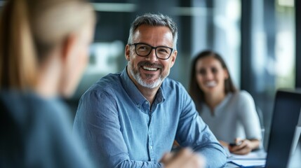 A smiling man in glasses engages in a conversation with two colleagues in a modern office setting, fostering a collaborative atmosphere.