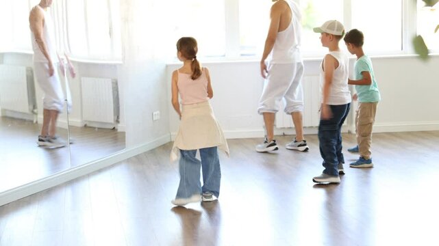 Preteen children enthusiastically following male instructor moves during group hip-hop lesson in sunlit mirrored studio, mastering modern street dance choreography. High quality 4k footage